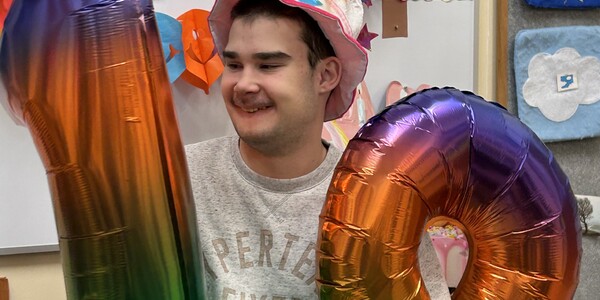Uczeń siedząc na krześle trzyma duże nadmuchane balony-cyfry: 1 i 9