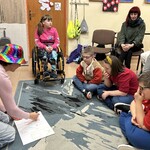 Uczniowie siedzą na podłodze i na krzesłach. Nauczyciel w kolorowym kapeluszu rysuje plan Wiaderka