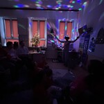 Nauczyciel trzyma w ręku papierową „racę” i zimne ognie. W pomieszczeniu jest ciemno, świecą się kolorowe lampki.