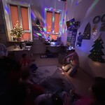 Nauczyciel trzyma w ręku papierową „racę” i zimne ognie. W pomieszczeniu jest ciemno, świecą się kolorowe lampki.