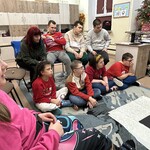 Uczniowie siedzą na podłodze i na krzesłach.