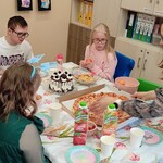 Uczniowie siedzą wokół stołu. Nauczycielka stawia pizzę na stole