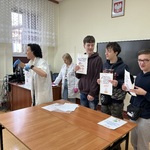 Uczniowie trzymają plansze z zasadami, które należy przestrzegać w trakcie eksperymentów. Nauczyciel stoi obok, tłumaczy.