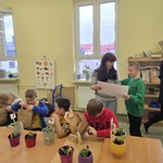 Uczniowie siedzą przy stole na którym stoją doniczki z posadzonymi kwiatami. Z tyłu uczeń wręcza Pani kartkę z namalowana praca plastyczną