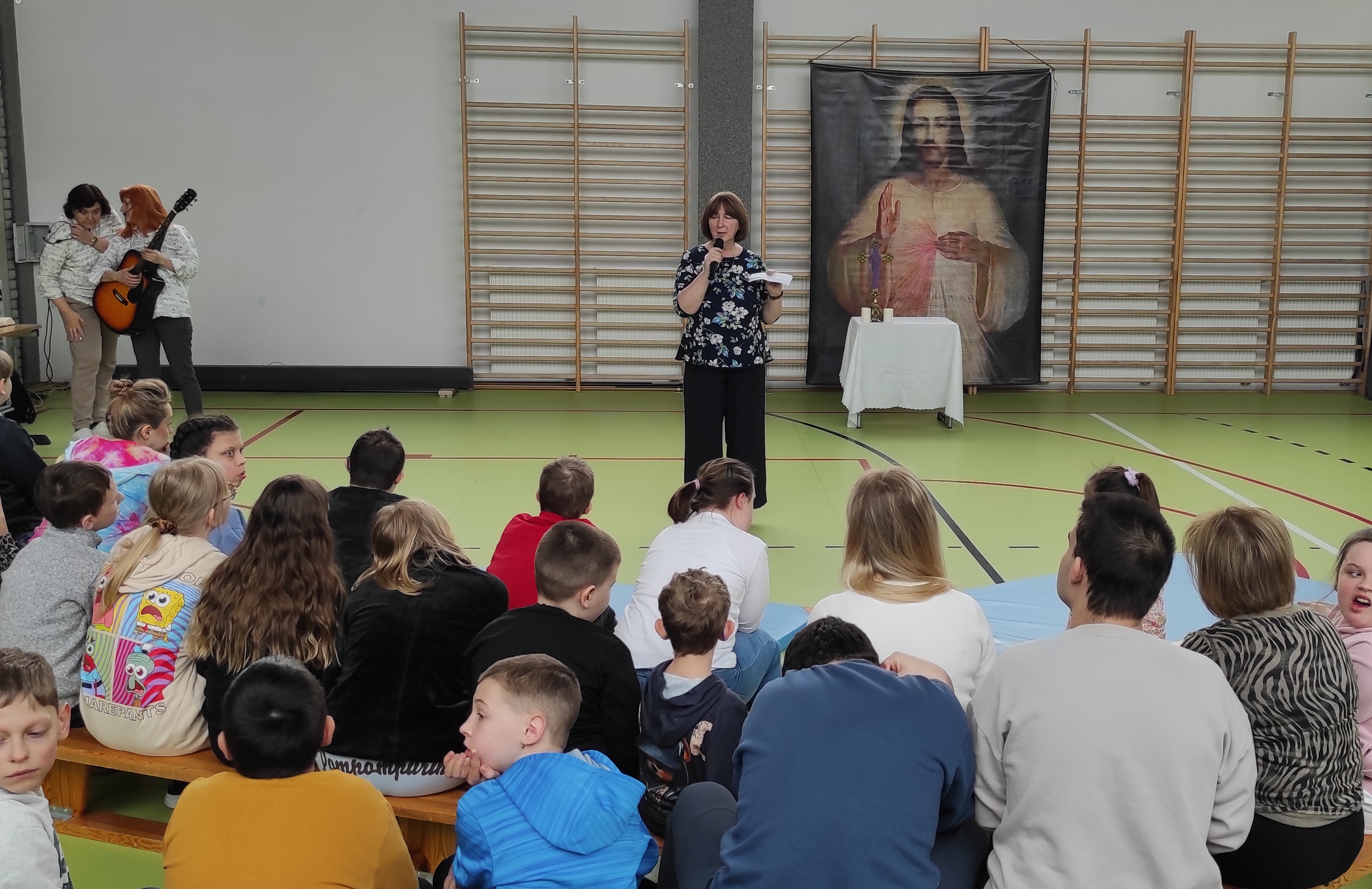 Nauczycielka mówi do uczniów siedzących na łąweczkach