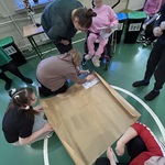 Nauczycielka pochylona nad uczennicą przyklejającą kartkę na duży arkusz szarego papieru.