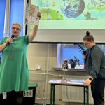 Jedna nauczycielka mówi do mikrofonu i pokazuje obrazek.  Obok druga nauczycielka z mikrofonem w dłoni  patrzy na stól