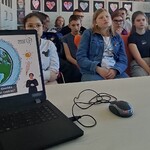 Laptop z wyświetlanym slajdem. Z tyłu uczniowie biorący udział w lekcji