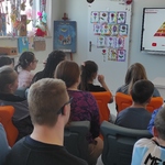 Uczniowie uczestniczący w lekcji patrzą na nauczycielkę omawiającą slajd wyświetlany na tablicy