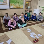 Grupa uczniów siedzi na podłodze. Przed nimi leżą arkusze papieru szarego