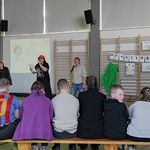Uczniowie siedzą na ławce. Trzy nauczycielki mówią do mikrofonów