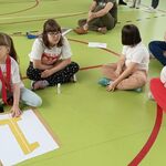 Grupa uczniów siedzi na podłodze na sali gimnastycznej. Przed nimi leży kartka z napisem 1