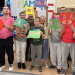 Uczniowie trzymają w ręku prace z ozdobnym napisem: mama.