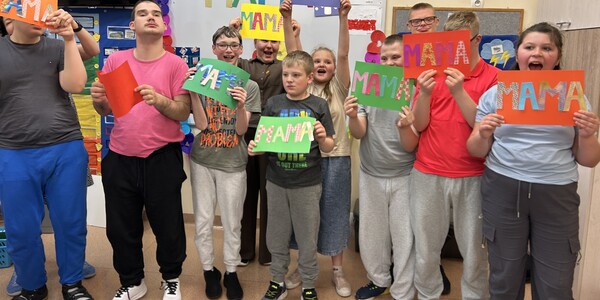 Uczniowie trzymają w ręku prace z ozdobnym napisem: mama.