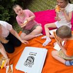 Pomoc i trójka uczniów siedzą na kocu i jedzą pizzę