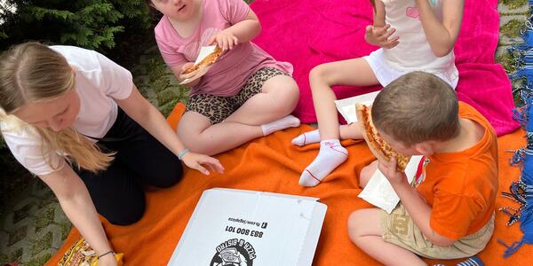 Pomoc i trójka uczniów siedzą na kocu i jedzą pizzę