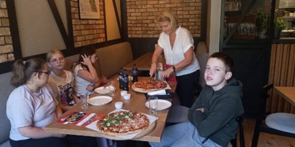 Nauczycielka kroi pizzę. Uczniowie siedzą przy stole w pizzerni