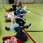 Grupa uczniów na sali gimnastycznej wykonuje ćwiczenie leżąc na plecach