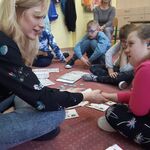 Nauczycielka i uczniowie siedzą na dywanie. Przed uczniami leżą książki do komunikacji. Nauczycielka patrzy na uczennicę i trzyma ją za rękę.