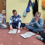 Uczniowie siedzą na dywanie. Przed uczniami leżą dwie książki komunikacyjne. Uczeń siedzący w środku pokazuje symbol w książce komunikacyjnej leżącej przed nim.