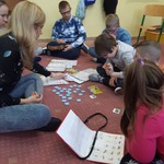 Nauczycielka i uczniowie siedzą na dywanie. Przed nimi leżą książki do komunikacji. Nauczycielka wskazuje symbol w książce do komunikacji uczennicy siedzącej w środku