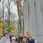 Pięcioro uczniów stoi razem z panią w okularach na tle wysokiego szarego Pomnika Żołnierza Polskiego