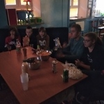 Grupa uczniów siedzi na kanapach. Przed nim stoi stół na którym znajdują się butelki z piciem i miski z popcornem.