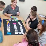 Grupa uczniów z nauczycielką siedzi wokół stołu na którym leży tablica z symbolami. Uczniowie przyglądają się