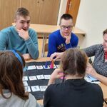 Grupa uczniów i nauczyciel siedzą wokół stołu na którym leży tablica  z symbolami.