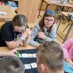 Uczniowie siedzący wokół stołu patrzą na zielony stożkowy pionek przesuwany na planszy przez widoczną na zdjęciu rękę nauczyciela.