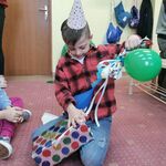 Uczeń w urodzinowej czapeczce siedzi na dywanie i rozpakowuje prezent opakowany w papier w kolorowe koła, wstążki i zielony balonik.