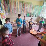 Nauczycielka trzyma tort przed uczniem na którym płonie świeczka. Obok stoją uczniowie z lurkami. Nad  uczniami wisi kolorowa dekoracja z pasków bibuły.