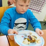 Chłopiec siedzi przy stole. Przed nim stoi talerz  z pokrojonymi na kawałki owocami. W prawej ręce trzyma widelec.