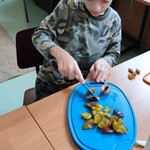 Uczeń siedzi przy stole i kroi śliwkę nożem na niebieskiej desce do krojenia.