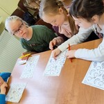 Grupa uczniów siedzi przy stole. Przed nimi leżą karty pracy. Uczennica w białej bluzce pokazuje właściwy obrazek koleżance siedzącej obok.