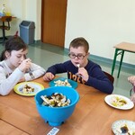 Czwórka uczniów siedzi przy stole. Przed nimi stoją talerze z potrawą. Na środku stołu stoi niebieska plastikowa miska z sałatką. Uczniowie trzymają widelce w dłoniach.