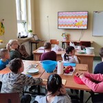 Uczniowie siedzą wokół stołu. Patrzą na kolorowy ekran telewizora, który wisi na ścianie. Na stole stoją miski, talerze, ręcznik papierowy, leżą deski do krojenia owoce i noże.