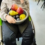 Dziewczynka siedzi na wózku inwalidzkim. Na kolanach trzyma metalową miskę z owocami.