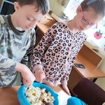 Dziewczynka i chłopiec stoją przy stole i mieszają  łyżkami kawałki owoców w niebieskiej misce.