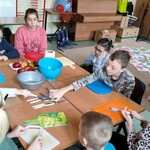 Grupa uczniów siedzi wokół stołu. Przed uczniami leżą kolorowe deski do krojenia. Na stole leżą noże  oraz stoją dwie plastikowe miski. Za miskami leżą czerwone owoce.