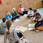 Uczniowie siedzą na dywanie po turecku w kółku. Przed nimi na dywanie leżą kartki z namalowanymi kolorowymi owocami.