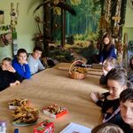 Grupa uczniów siedzi wokół stołu. Na stole leżą liście i stoi koszyk z liśćmi. Na ścianie znajduje się foto makieta z drzewami.