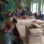 Uczniowie siedzą wokół dużego stołu. Przed nimi na stole stoi pudło kartonowe, małe pudełko oraz koszyk pełne suchych liści. U szczytu stołu stoi kobieta. Za nią znajduje się fototapeta - drzewa.