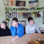 Dwóch chłopców i dziewczyna siedzą przy stole. Przed nimi stoi kosz pełen liści. Za nimi na ścianie  na sznurkach wiszą zdjęcia dzikach zwierząt.