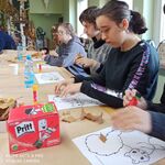 Uczniowie siedzą przy stole. Przed nimi leżą kartki z szablonem wiewiórki, liście, stoi pudełko z klejem oraz pojedyncze kleje i butelki z wodą.