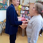 Chłopak i dziewczyna stoją bokiem i trzymają się za rękę. Z tyłu na tle regału z książkami przy stole siedzą dwie dziewczyny.