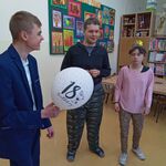 Dwóch chłopców i dziewczyna stoją na tle kolorowej gazetki oraz regału. Uczeń z przodu trzyma biały balon z napisem 18.