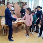 Dziewczyna trzyma za rękę chłopaka w garniturze. Z tyłu stoi grupa uczniów i się im przygląda.