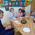 Grupa uczniów siedzi przy stole przykrytym kraciastą serwetą. Na stole stoją talerze i ciasto. Nad stołem wisi złoty balon