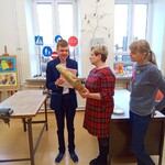 Dwie panie stoją obok chłopaka w niebieskim garniturze. Pani w sukience w kratkę trzyma prezent.
